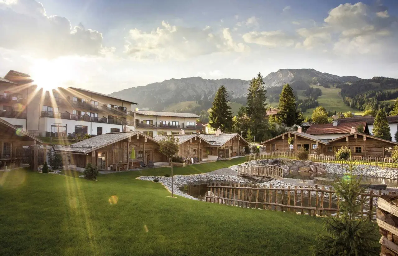 Alpin Chalets Oberjoch: Idyllische Hotelanlage mit traditionellen Chalets, Teich und majestätischem Alpenpanorama im Abendlicht.
