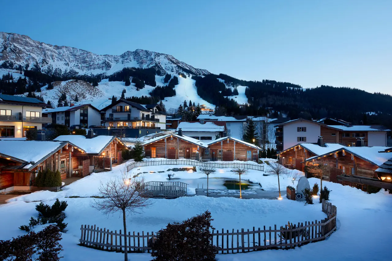 Alpin Chalets Oberjoch im Winter bei Dämmerung mit verschneiten Holzhäusern, beleuchtetem Teich und Skipiste im Hintergrund.