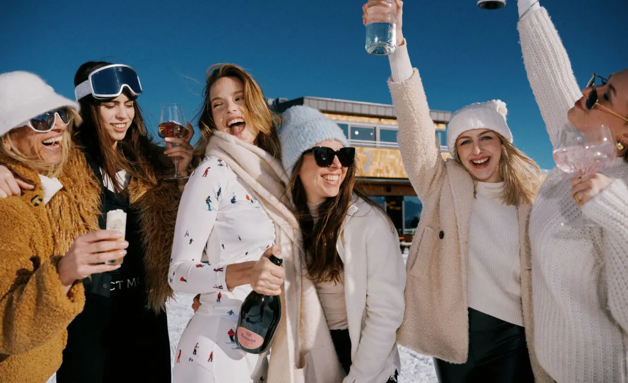 Lachende Frauen genießen Après-Ski mit Sekt und Drinks im Schnee vor dem Hotel Nesslerhof in den österreichischen Alpen.