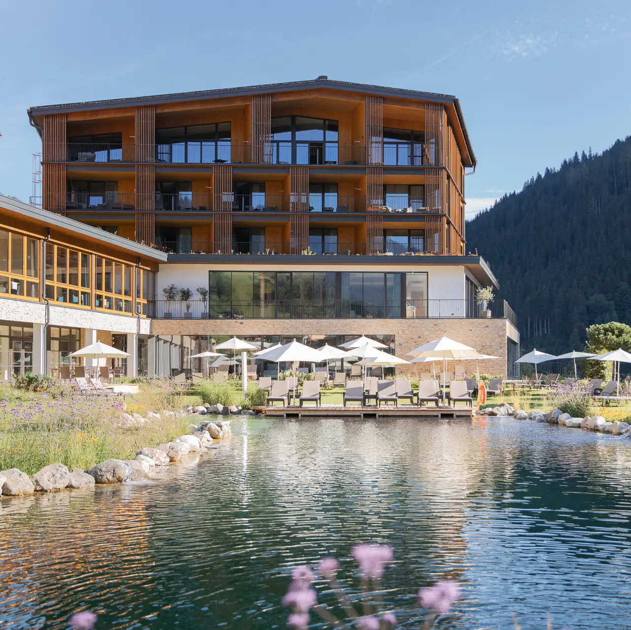 Natürlicher Badeteich mit Liegewiese und modernem Hotel Nesslerhof im Hintergrund, umgeben von Alpenpanorama.