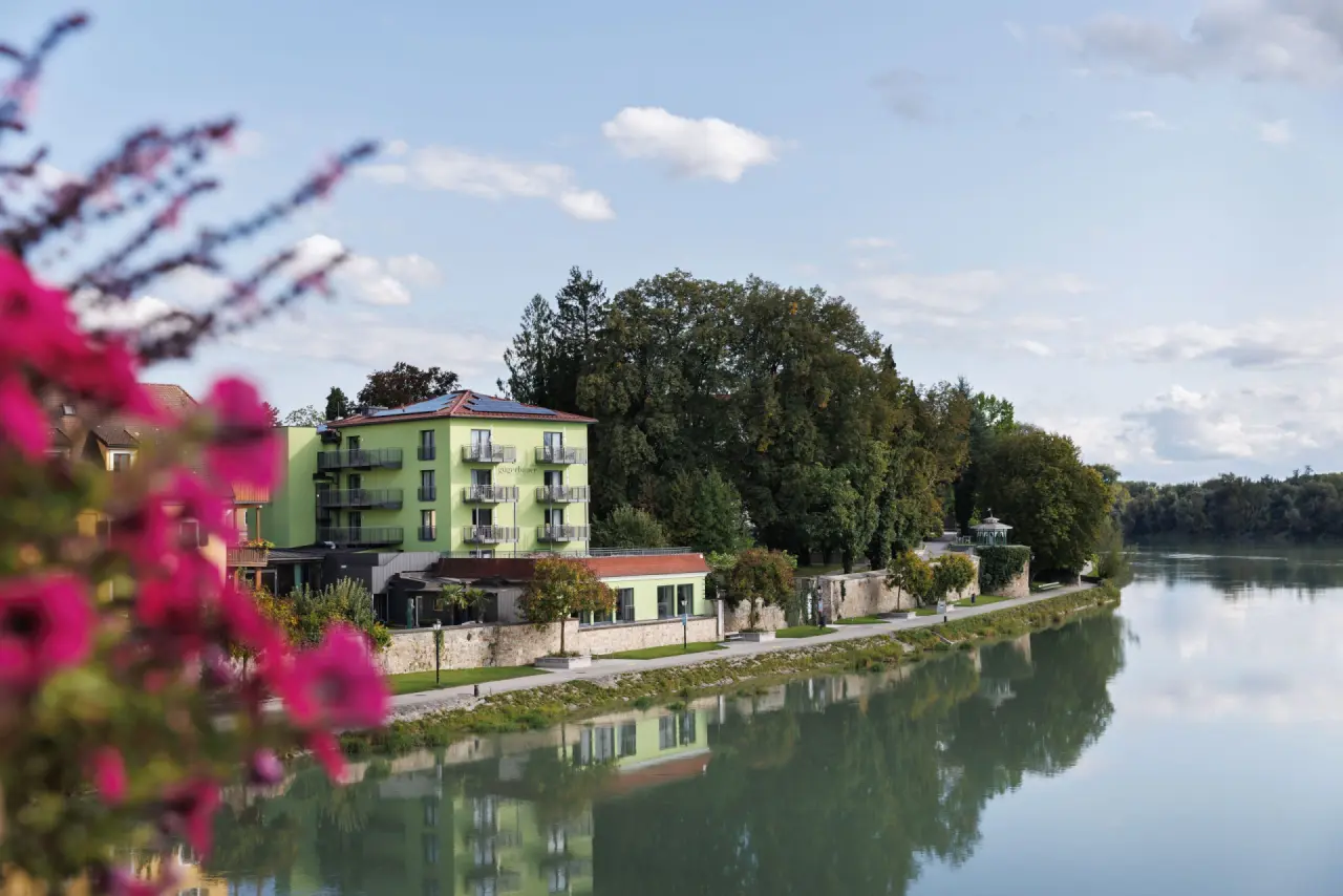 Hotel Gugerbauer am Flussufer mit grüner Fassade, Balkonen und Bäumen, gespiegelt im Wasser. Idyllische Lage.