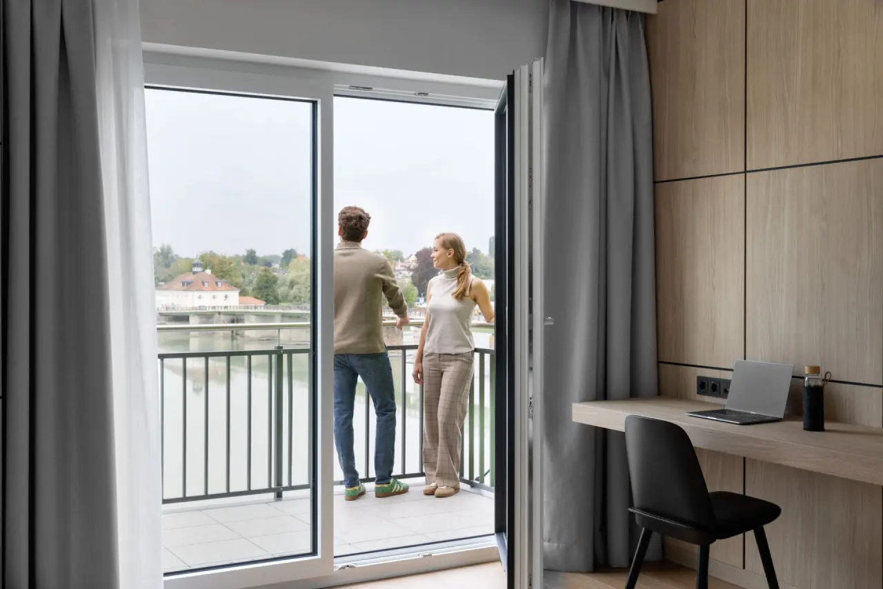 Paar genießt den Ausblick vom Balkon eines modernen Hotelzimmers im Hotel Gugerbauer mit Fluss- und Stadtpanorama.