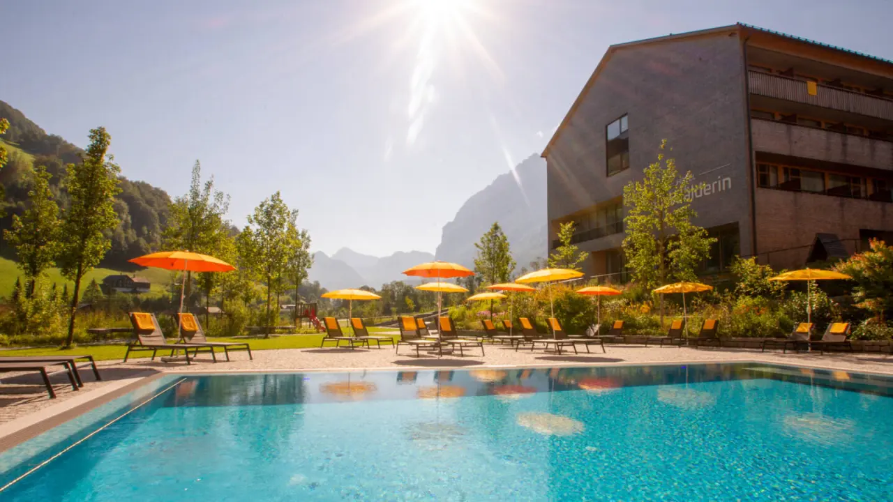 Aussenpool mit Liegestühlen und Sonnenschirmen vor der Bergkulisse des Hotel die Wälderin****s an einem sonnigen Tag.