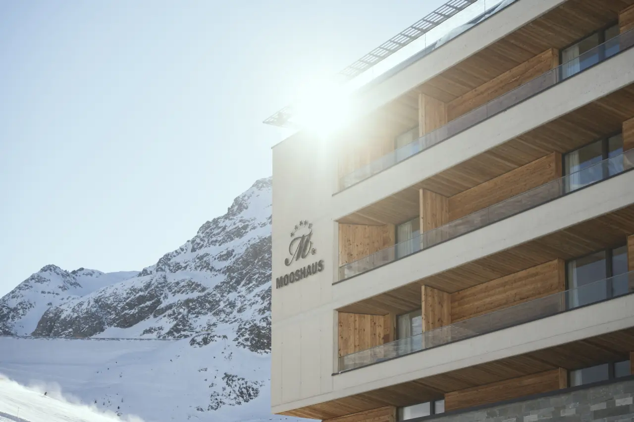 Modernes Hotel Mooshaus mit Holzbalkonen und Sonnenterrasse vor verschneiter Bergkulisse in den Alpen.