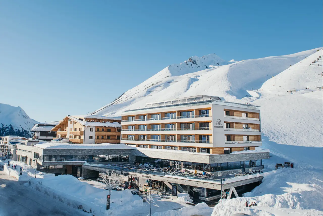 Panoramablick auf das moderne Hotel Mooshaus in den verschneiten Alpen mit Skigebiet und Sonnenterrasse.