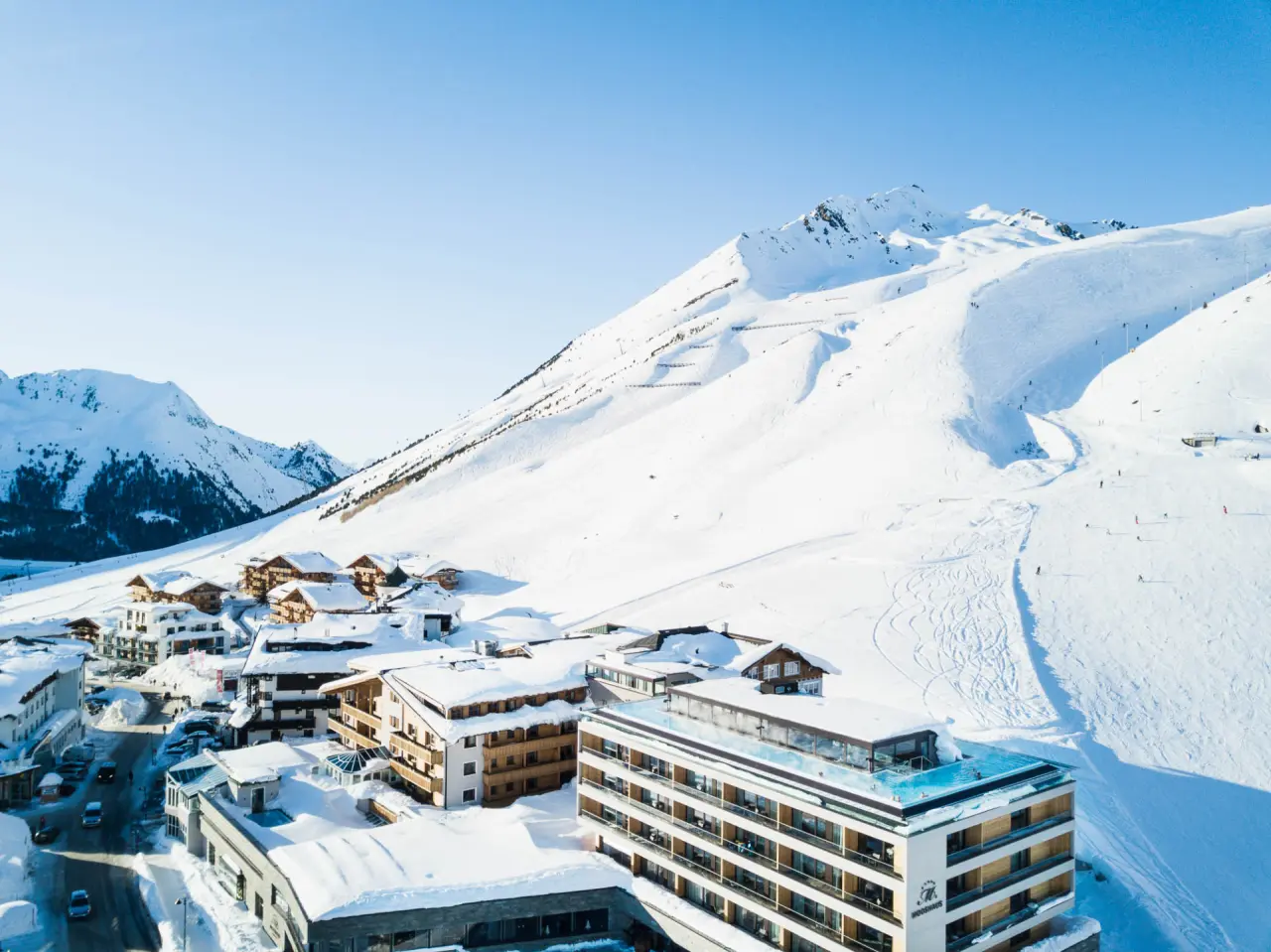 Luftaufnahme des Hotel Mooshaus mit Infinity Pool auf dem Dach und schneebedeckten Bergen im Skigebiet.