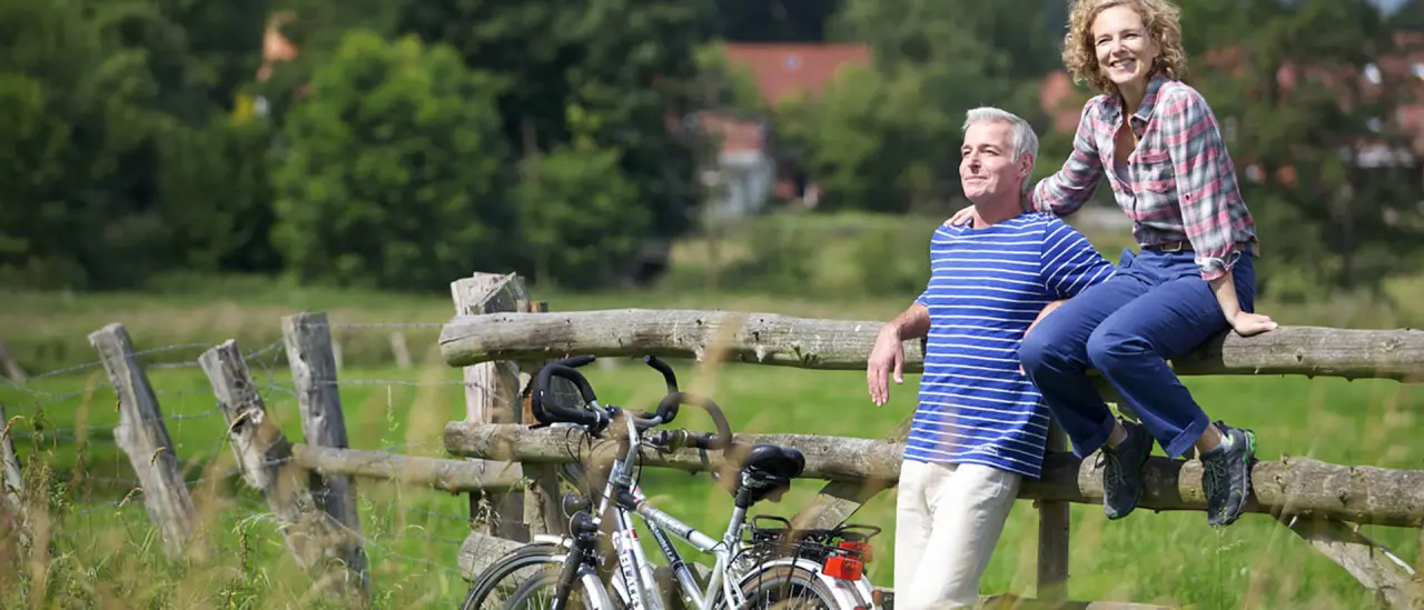 Ein lächelndes Paar macht eine Fahrradpause an einem Holzzaun in der grünen Natur, typisch für das Ringhotel Sellhorn.