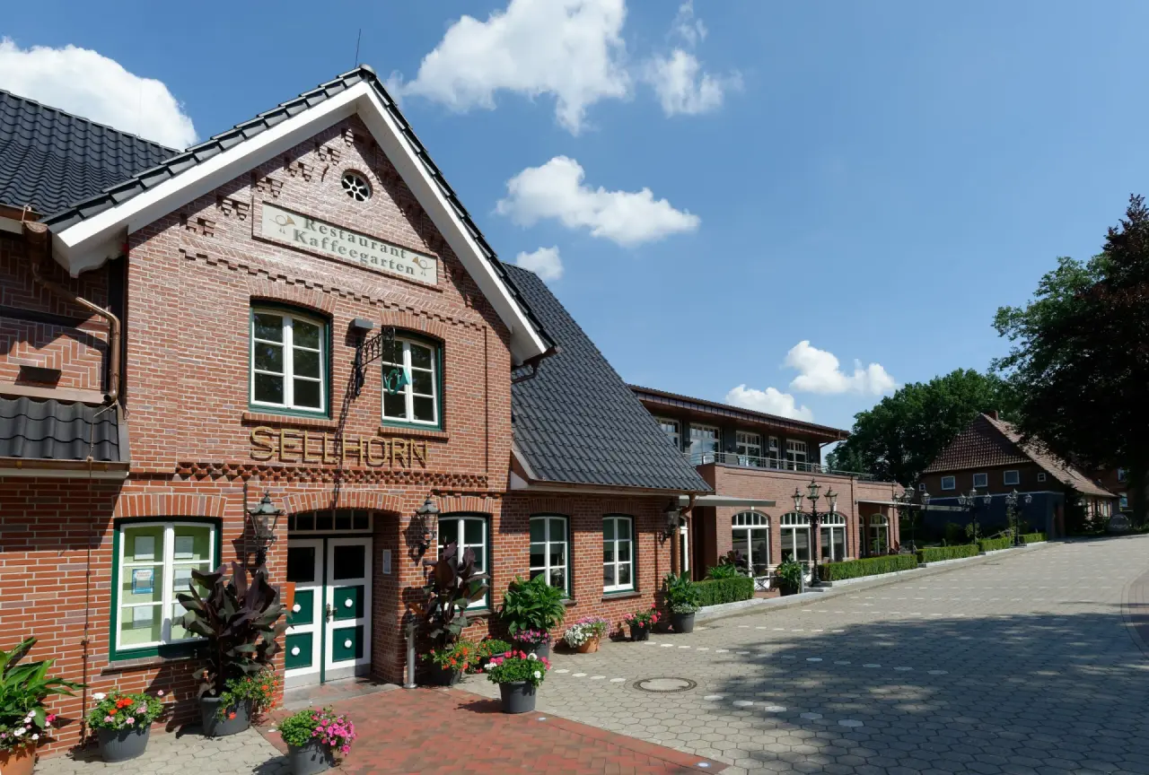 Aussenansicht des Ringhotel Sellhorn mit roter Backsteinfassade, dunklem Dach und blühenden Pflanzen unter blauem Himmel.