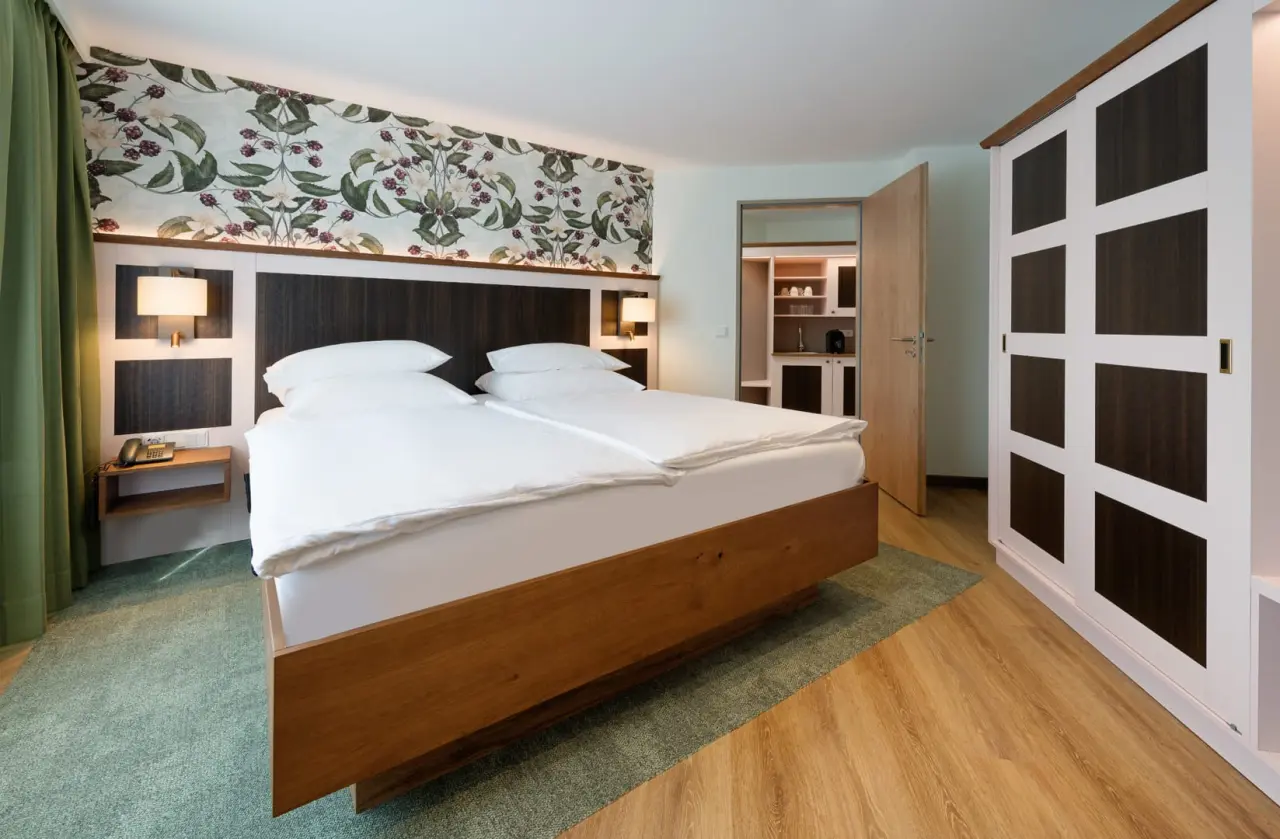 Gemütliches Doppelzimmer im Ringhotel Sellhorn mit floraler Tapete, Holzbett und kleiner Küchenzeile.