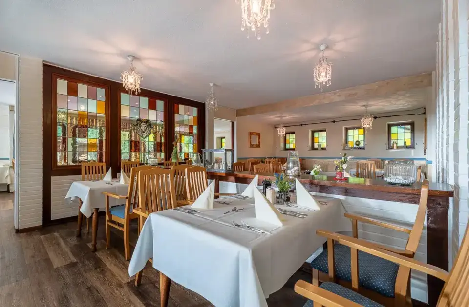Gemütlicher Restaurantbereich im Hotel Zur Heidschnucke mit festlich gedeckten Tischen und farbigen Bleiglasfenstern.