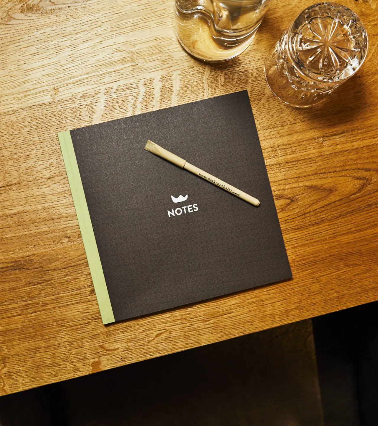 Notizbuch und Stift mit DAS KRONTHALER Logo auf Holztisch, daneben zwei Wassergläser. Detailaufnahme.