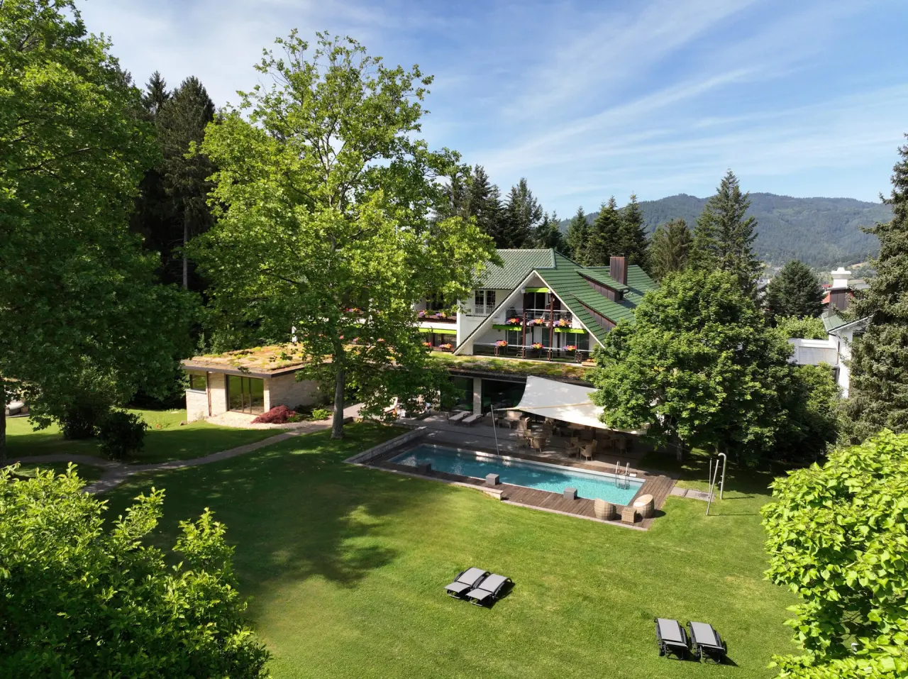 Aussenansicht des Waldhotel Grüner Baum mit Pool, Liegewiese und umgebendem Wald unter blauem Himmel.