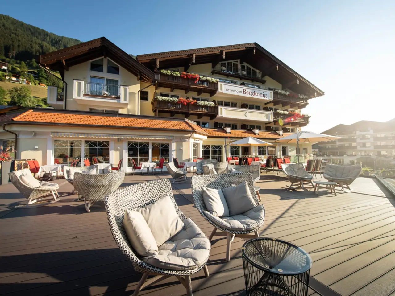 Aussenansicht des Activhotel Bergkönig mit sonniger Terrasse und modernen Lounge-Möbeln vor alpiner Bergkulisse.