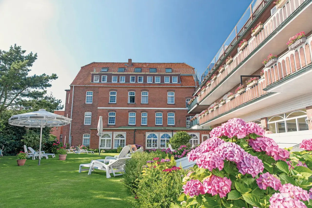 Aussenansicht des Nordseehotel Freese mit rotem Backsteingebäude, Balkonen und blühendem Garten mit Liegestühlen unter blauem Himmel.