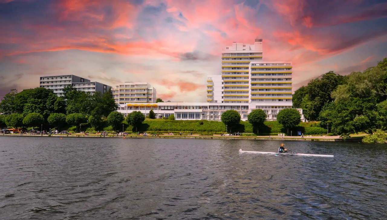Vitalia Seehotel Aussenansicht am Seeufer mit Ruderboot und dramatischem Sonnenuntergangshimmel.