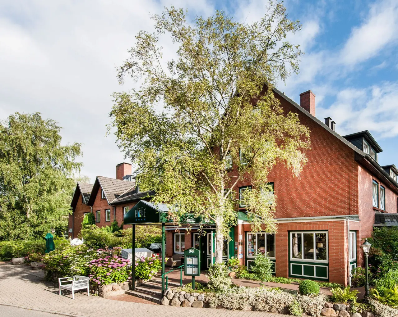 Aussenansicht des Hotel Birke mit roter Backsteinfassade, grünen Bäumen und blühenden Hortensien am Eingang.