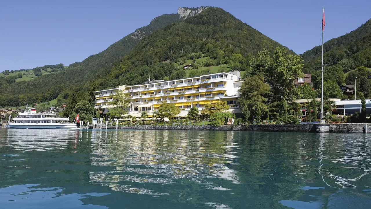 Panoramablick auf das BEATUS Wellness- & Spa-Hotel am Thunersee, umgeben von grünen Bergen, mit Schiff am Steg.