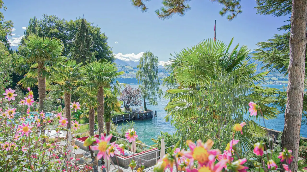 Blühender Hotelgarten mit Palmen und Terrasse, Blick auf den Thunersee und die Alpen im BEATUS Wellness- & Spa-Hotel.