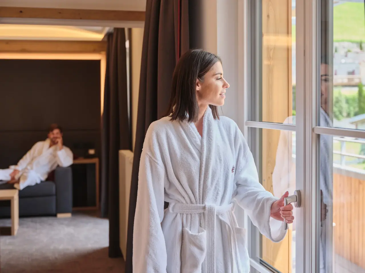 Entspanntes Paar in Bademänteln im komfortablen Hotelzimmer des Sporthotel Wagrain mit Blick ins Grüne.