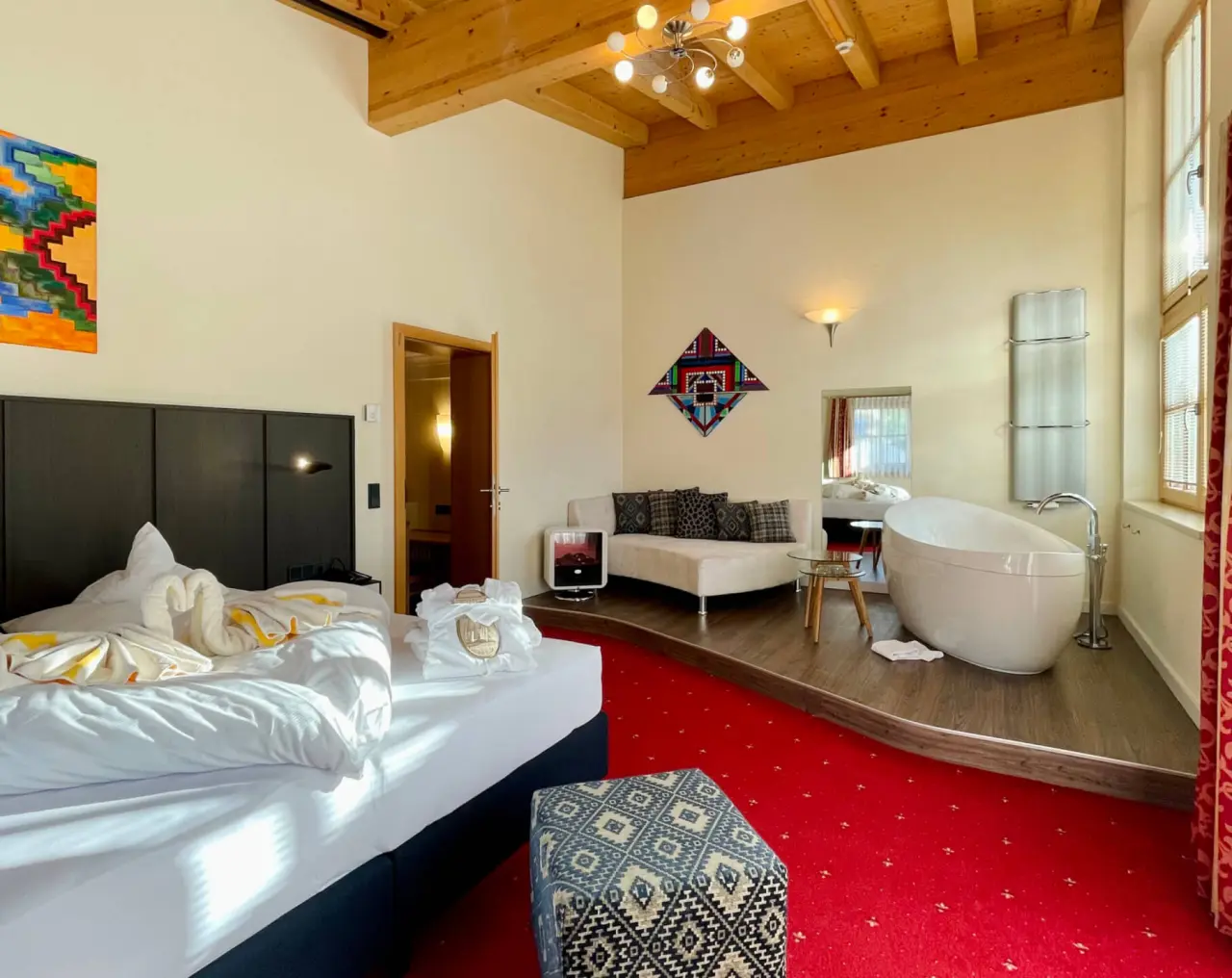 Geräumiges Hotelzimmer im Wellness- & Familienhotel Egger mit freistehender Badewanne, Holzbalken und rotem Teppich.