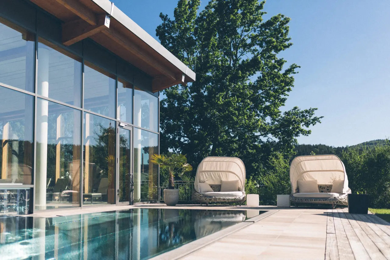 Aussenpool mit modernen Liegen und Glasfassade des Wellnessbereichs im Hotel Eibl-Brunner, umgeben von grüner Natur.
