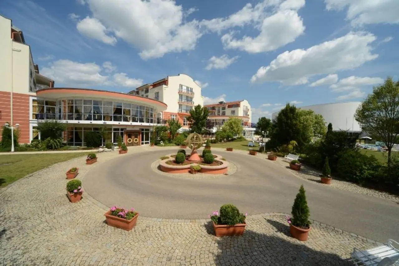 Aussenansicht des The Monarch Hotel mit Haupteingang, gepflegter Zufahrt und Andeutung des Aussenpools unter blauem Himmel.