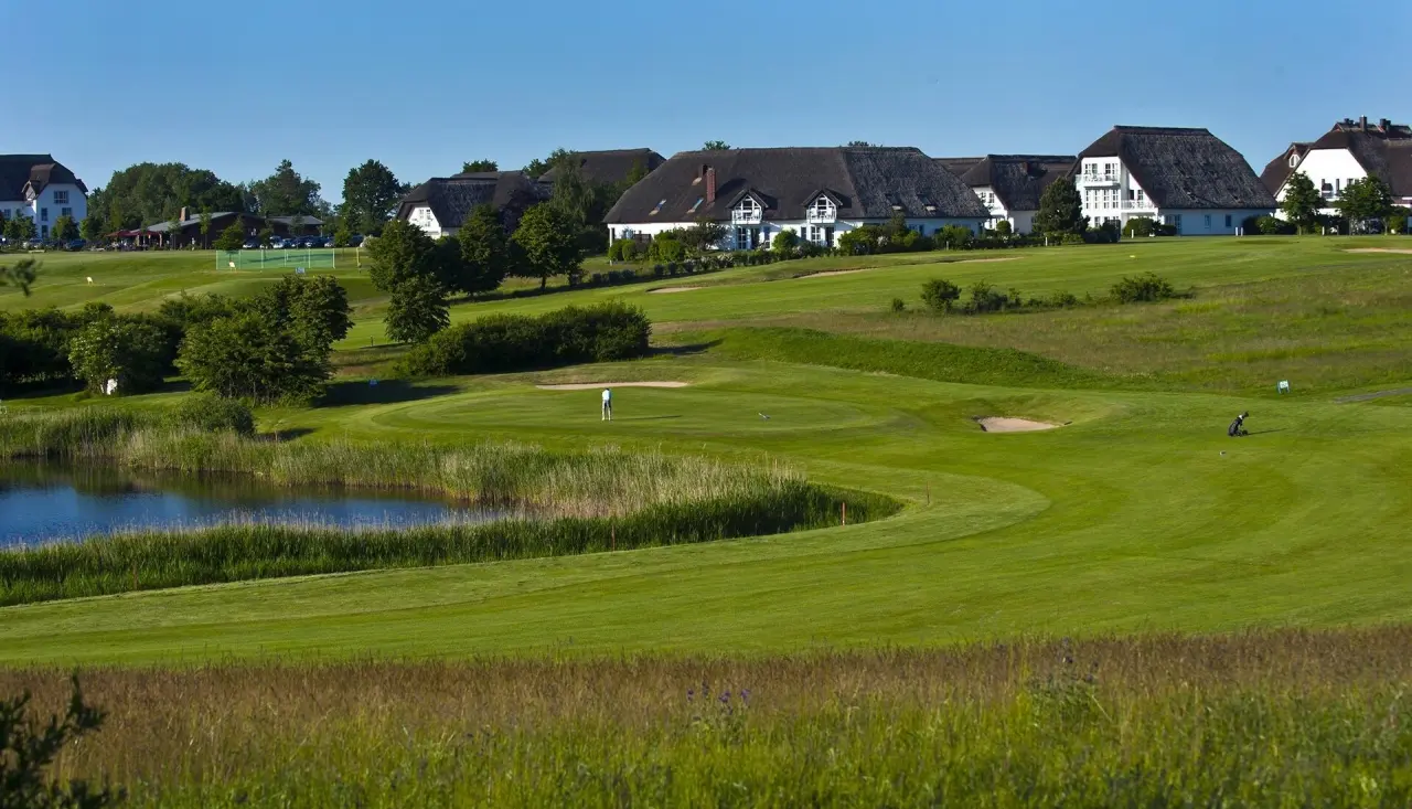 Weitläufiger Golfplatz mit idyllischem See und traditionellen Reetdachhäusern des Balmer See - Hotel·Golf·Spa unter blauem Himmel.