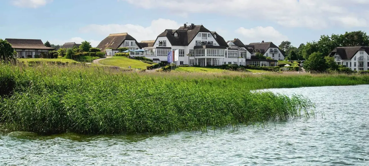 Aussenansicht des Balmer See - Hotel·Golf·Spa mit Reetdächern am Ufer des Sees unter blauem Himmel.