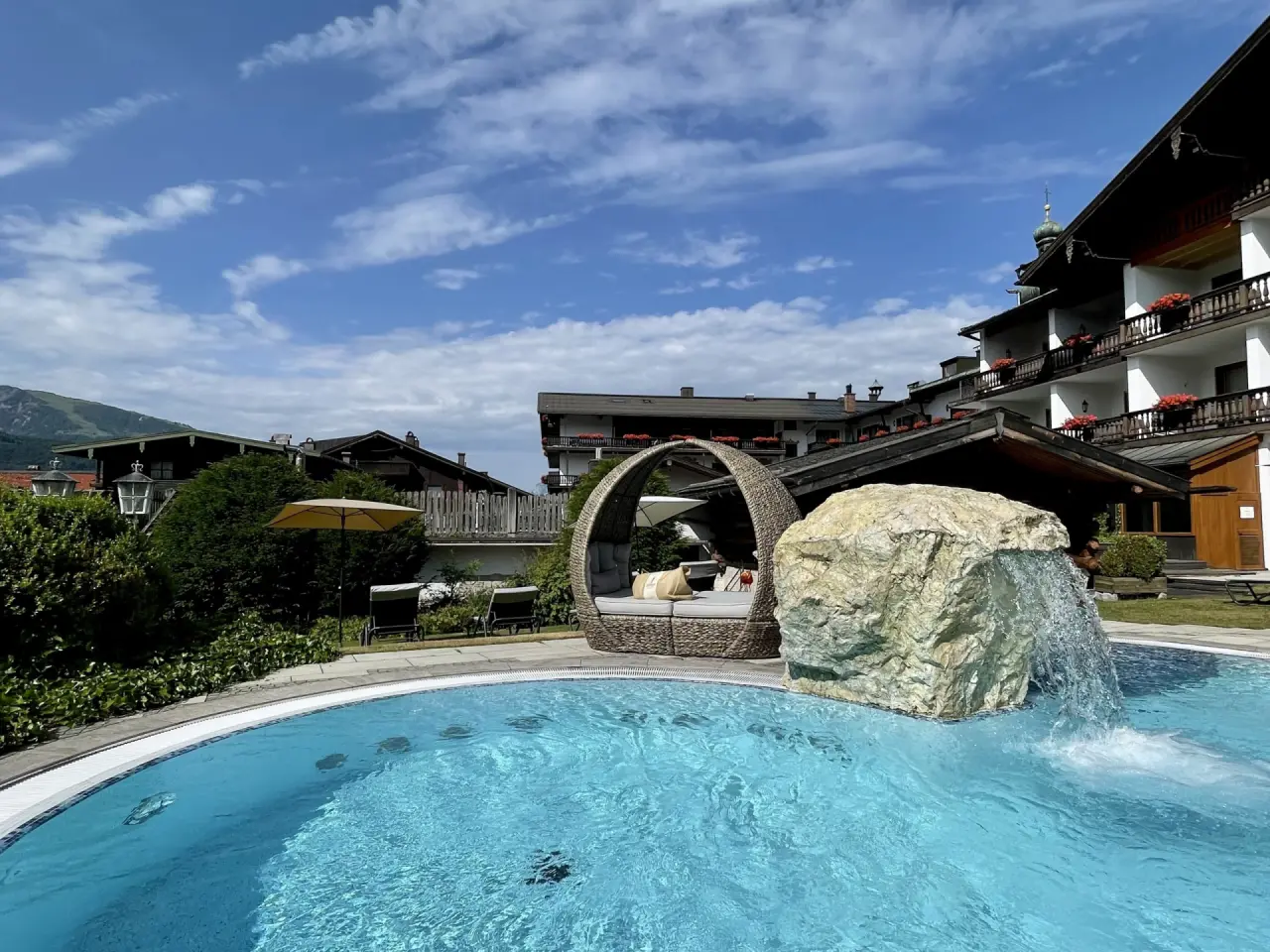 Aussenpool mit Wasserfall, Daybed und Liegen im Garten des Hotel Unterwirt unter blauem Himmel.