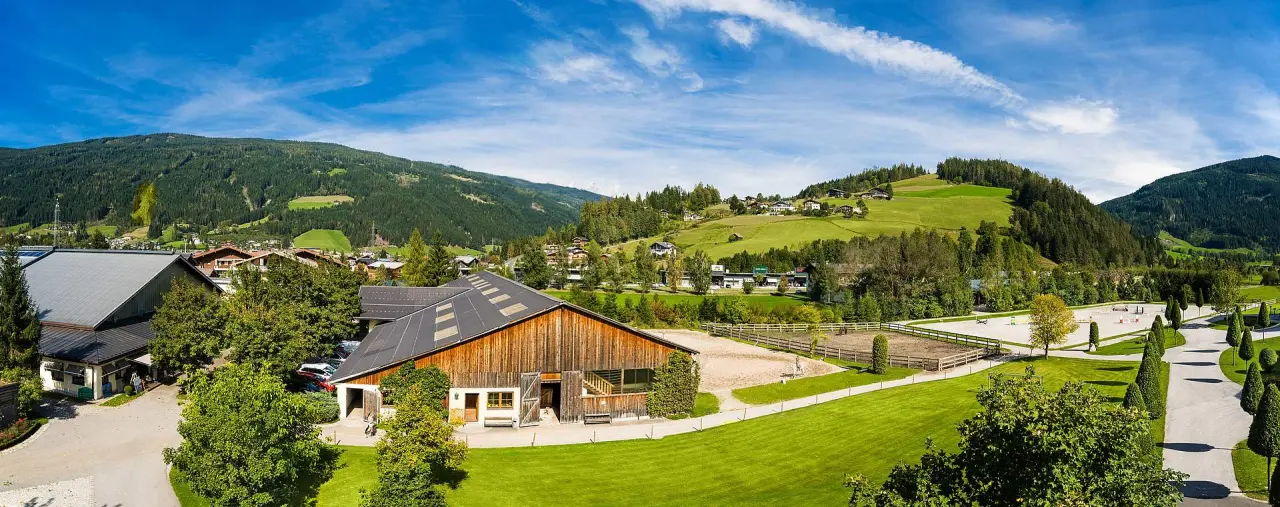Panoramablick auf Hotel Gut Weissenhof Radstadt mit Reitplatz, Stallungen und grüner Berglandschaft unter blauem Himmel.
