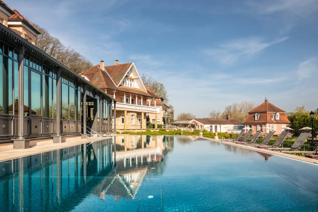 Infinity Pool und historische Gebäude des BollAnts Spa im Park unter blauem Himmel, einladende Liegebereiche am Wasser.