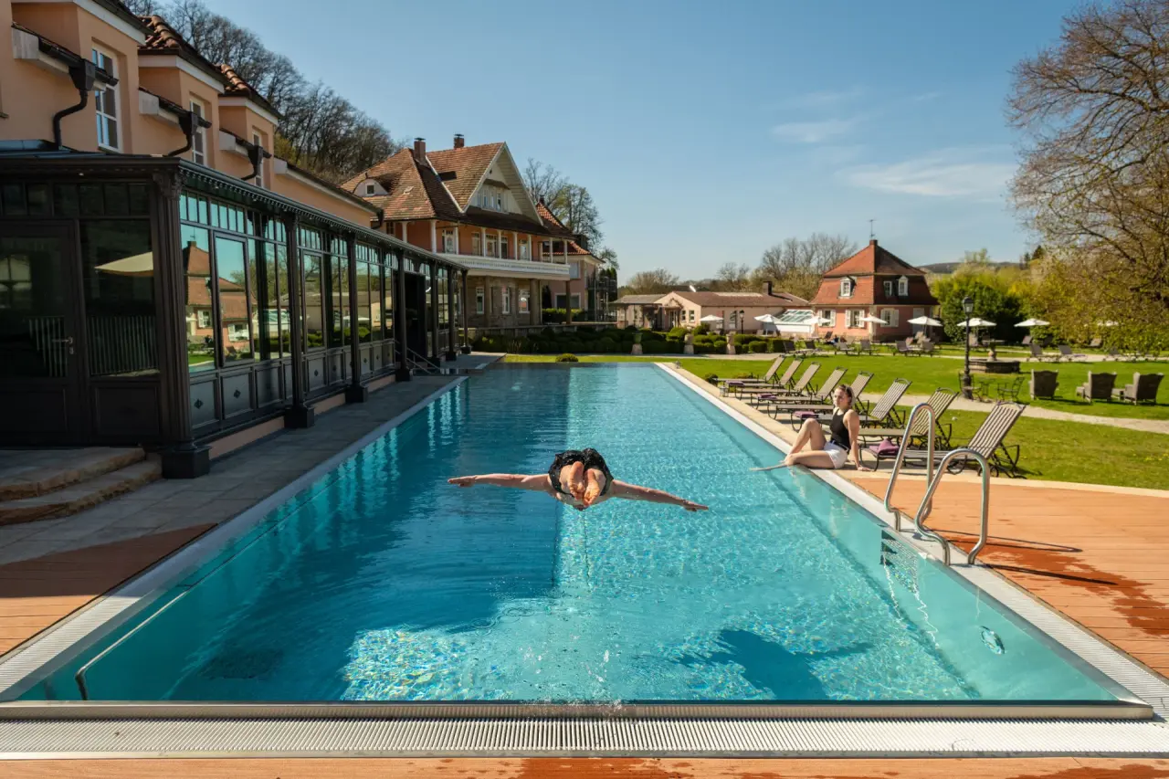 Aussenpool mit Infinity-Rand und sonniger Liegewiese im BollAnts Spa im Park. Eine Person taucht ins Wasser, eine andere entspannt am Beckenrand.