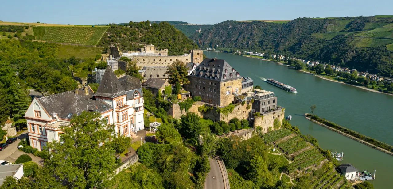 Panoramablick auf das historische Hotel Schloss Rheinfels am Rhein mit Weinbergen und vorbeifahrendem Schiff.