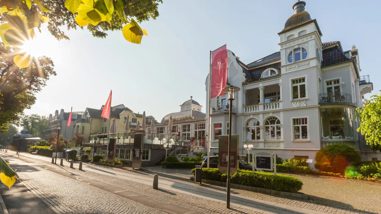 Sonnige Aussenansicht des Hotel Vier Jahreszeiten Kühlungsborn mit eleganter Bäderarchitektur und Kopfsteinpflaster.