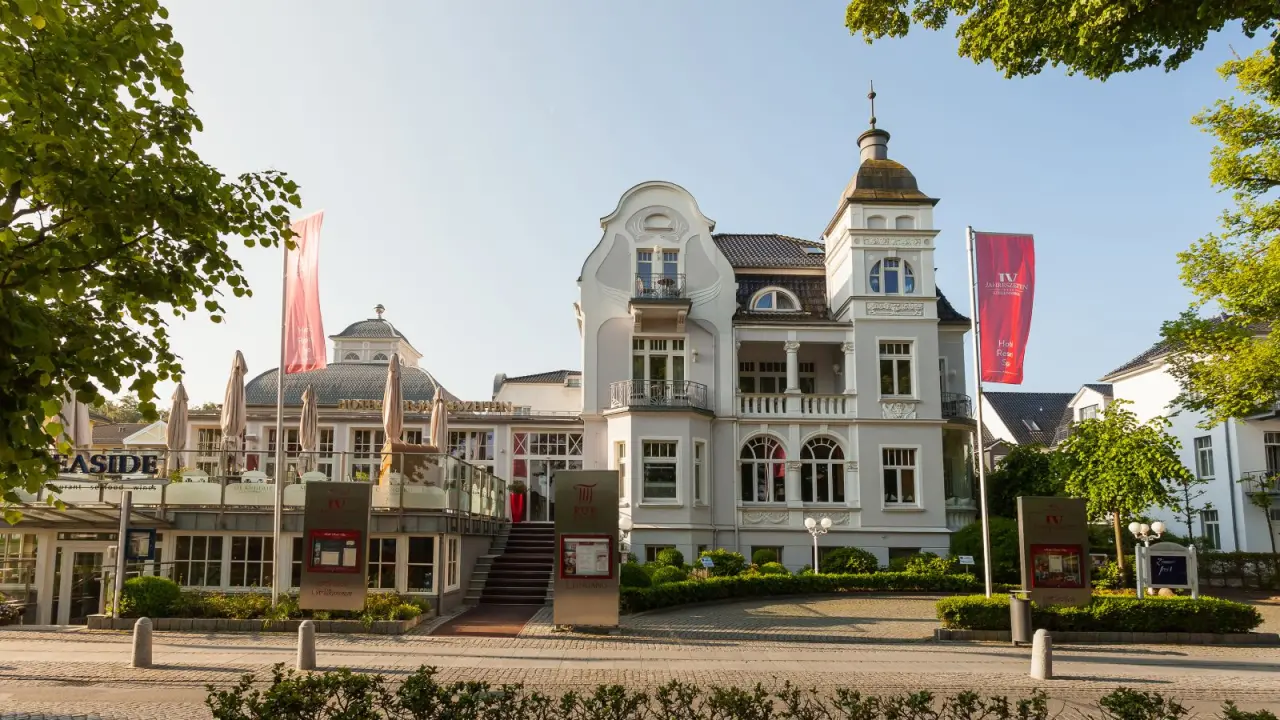 Elegante Aussenansicht des Hotel Vier Jahreszeiten Kühlungsborn mit historischer Architektur und grünen Bäumen unter blauem Himmel.