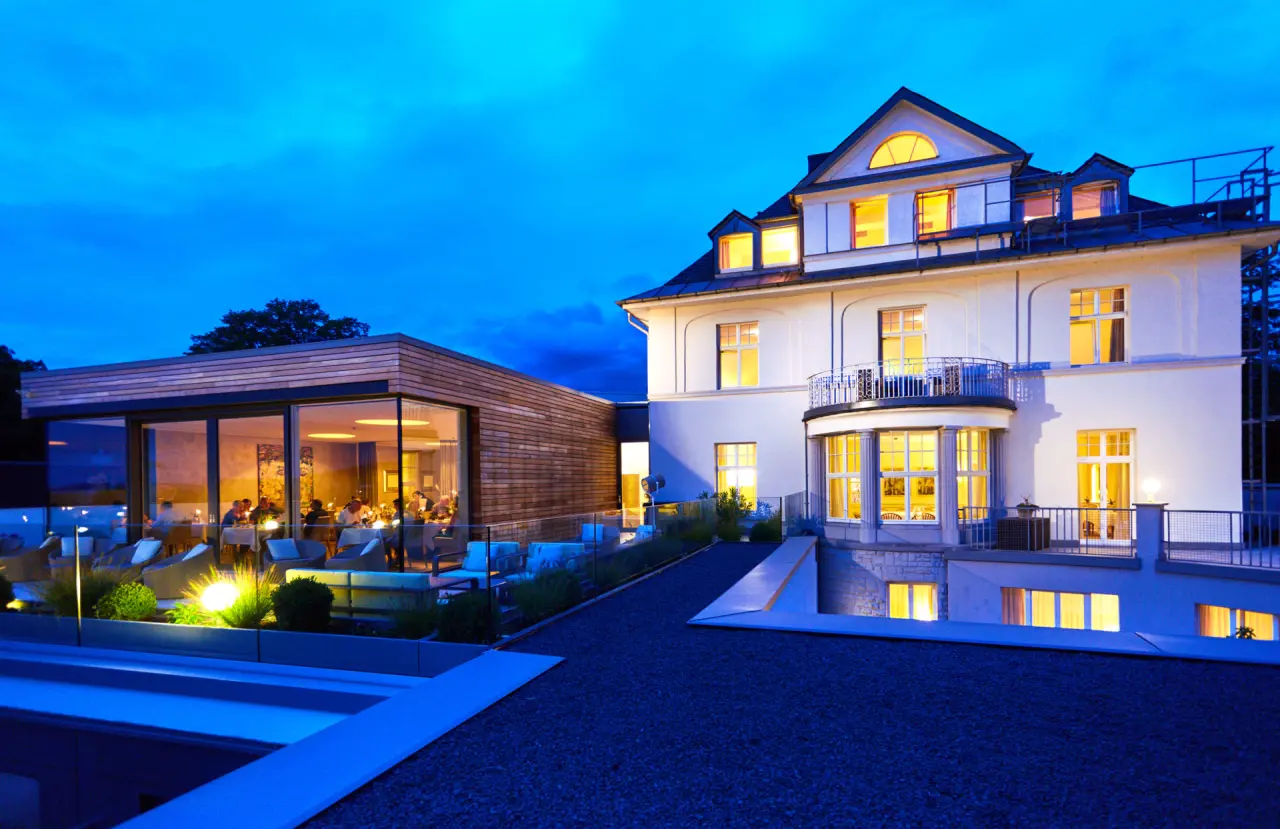 Beleuchtete Aussenansicht des eleganten Hotel Villa Hügel bei Dämmerung mit Restaurant und Terrasse.