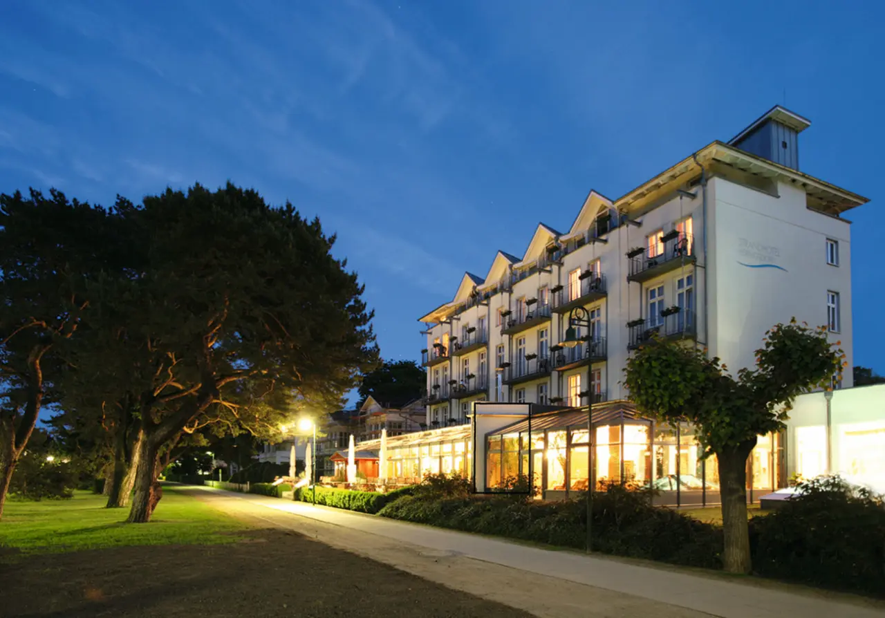 Abendansicht des Strandhotel Heringsdorf an der Ostsee mit beleuchteter Fassade und Restaurantterrasse unter blauem Himmel.
