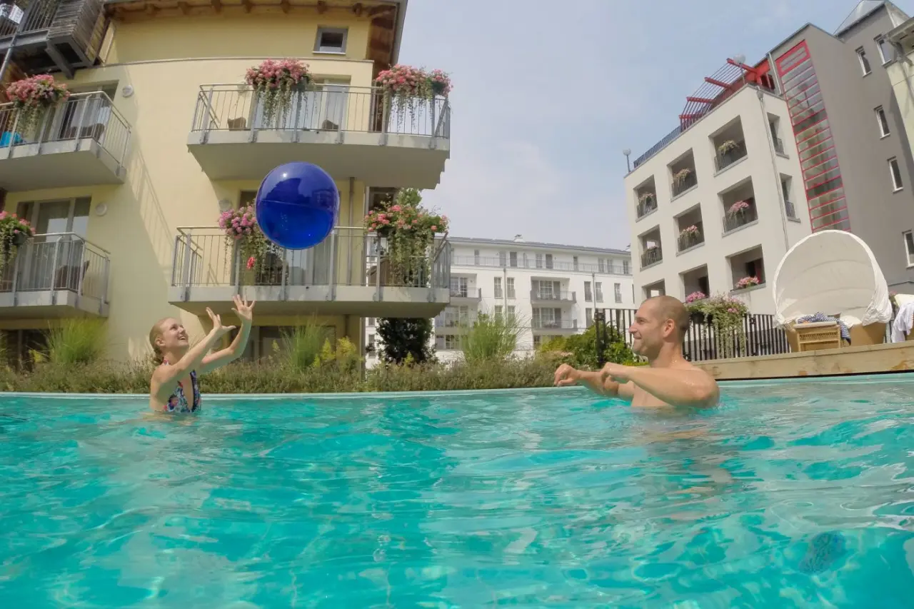 Gäste spielen vergnügt mit einem Wasserball im Aussenpool des Strandhotel Heringsdorf, umgeben von blühenden Balkonen.