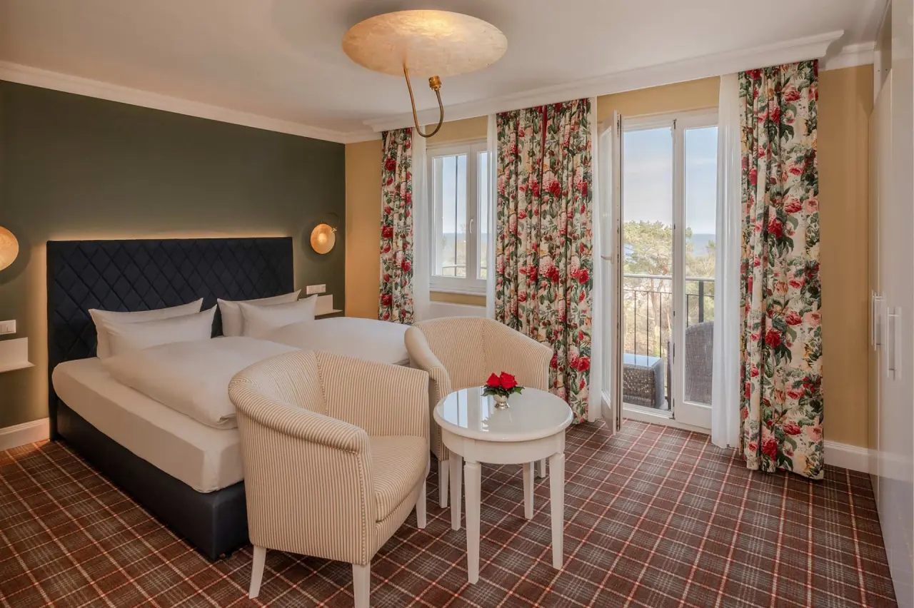 Helles Hotelzimmer im Strandhotel Heringsdorf mit Doppelbett, floralen Vorhängen und Balkon mit Meerblick.