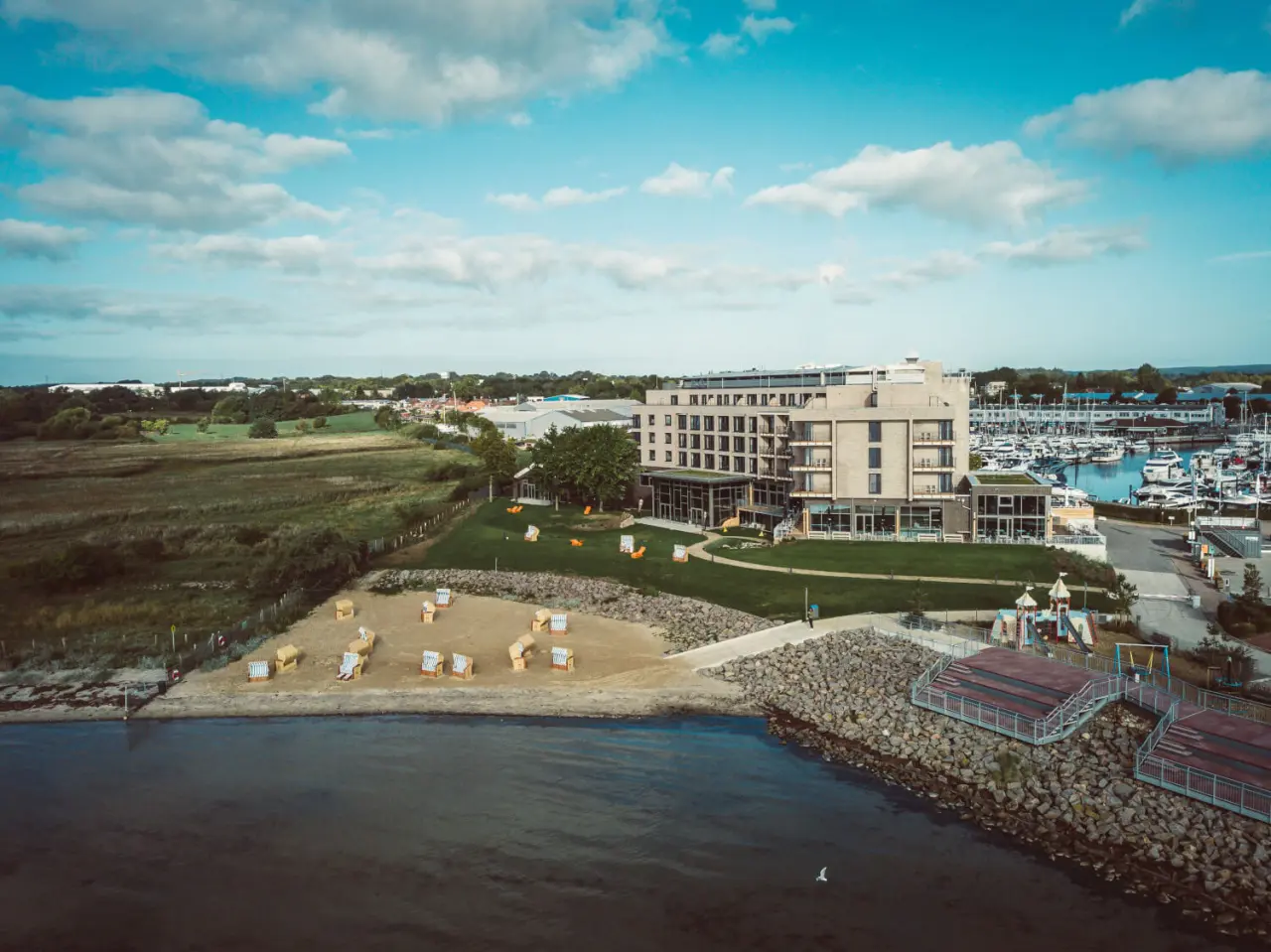 Luftaufnahme des ARBOREA Marina Resort Neustadt mit Hotelgebäude, Strand, Yachthafen und Ostsee unter blauem Himmel.