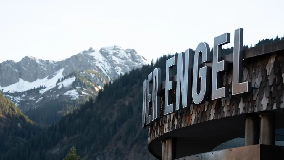 Schriftzug 'DER ENGEL' am Wellnesshotel Engel mit majestätischen schneebedeckten Alpen und grünen Wäldern im Hintergrund.