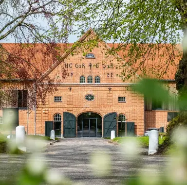 Der historische Eingang des Hotel Breitenburg, ein Backsteingebäude mit grünen Toren, umrahmt von frischem Frühlingslaub.