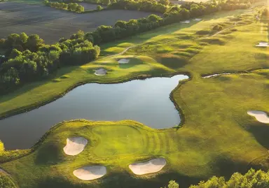 Luftaufnahme des weitläufigen Golfplatzes mit See und Bunkern am Hotel Breitenburg im Abendlicht.