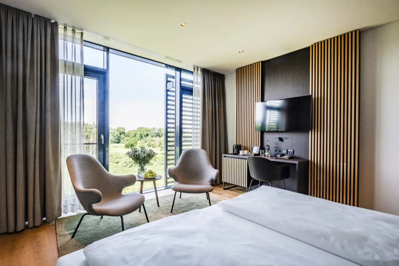 Modernes Hotelzimmer im Hotel Breitenburg mit bodentiefen Fenstern, gemütlicher Sitzecke und Blick ins Grüne.