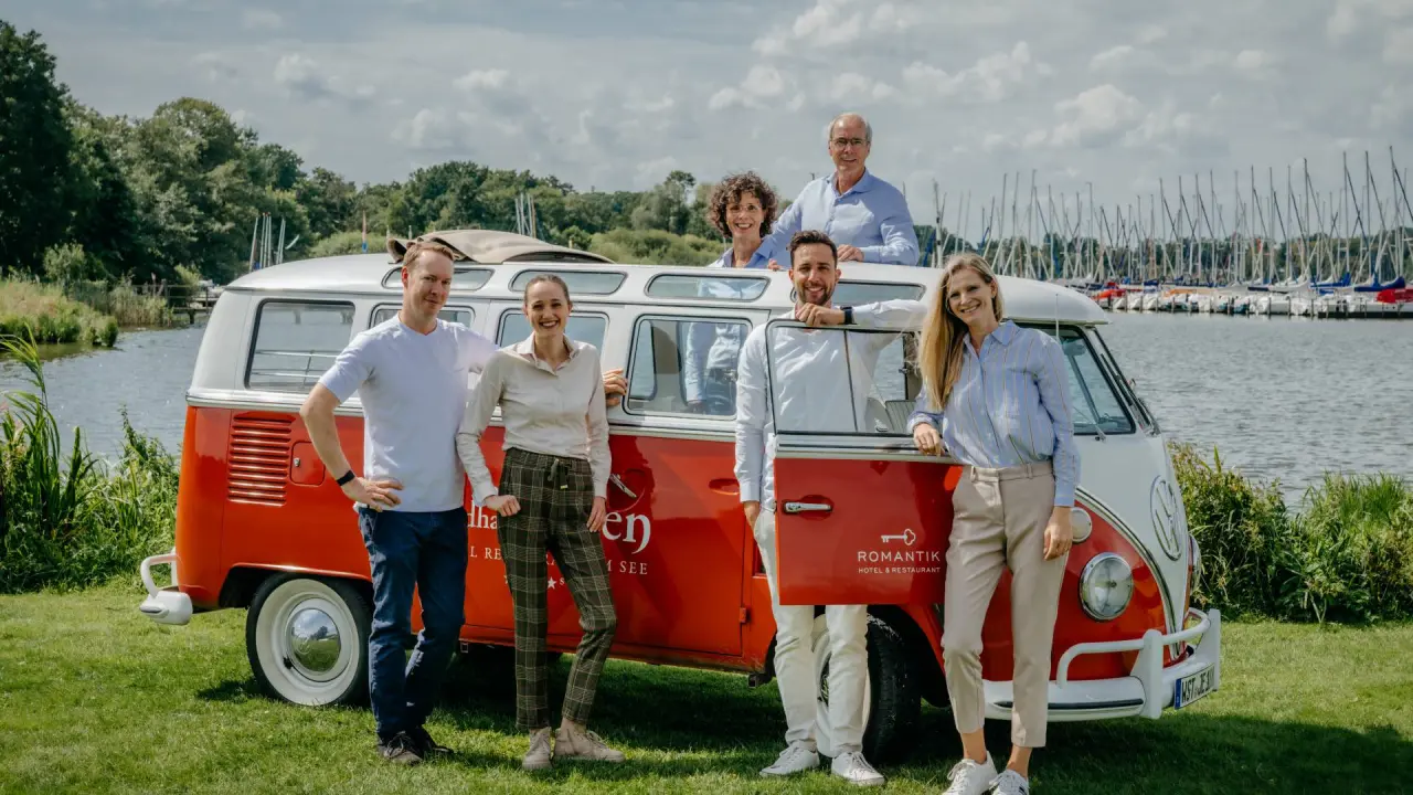 Team des Romantik Hotel Jagdhaus Eiden mit Oldtimer VW Bus am Zwischenahner Meer mit Segelbooten im Hintergrund.