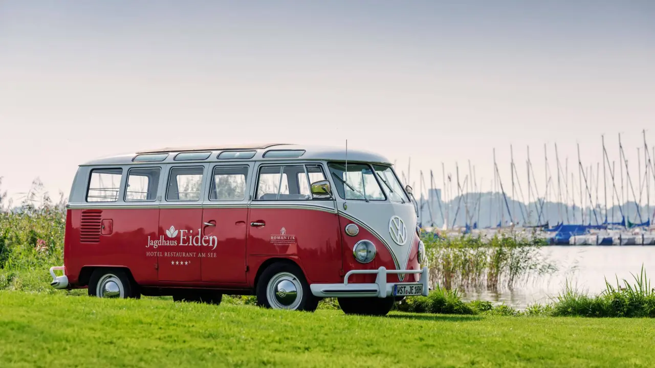 Roter Vintage VW Bus mit Jagdhaus Eiden Logo am Seeufer, im Hintergrund Segelboote. Romantik Hotel Jagdhaus Eiden.