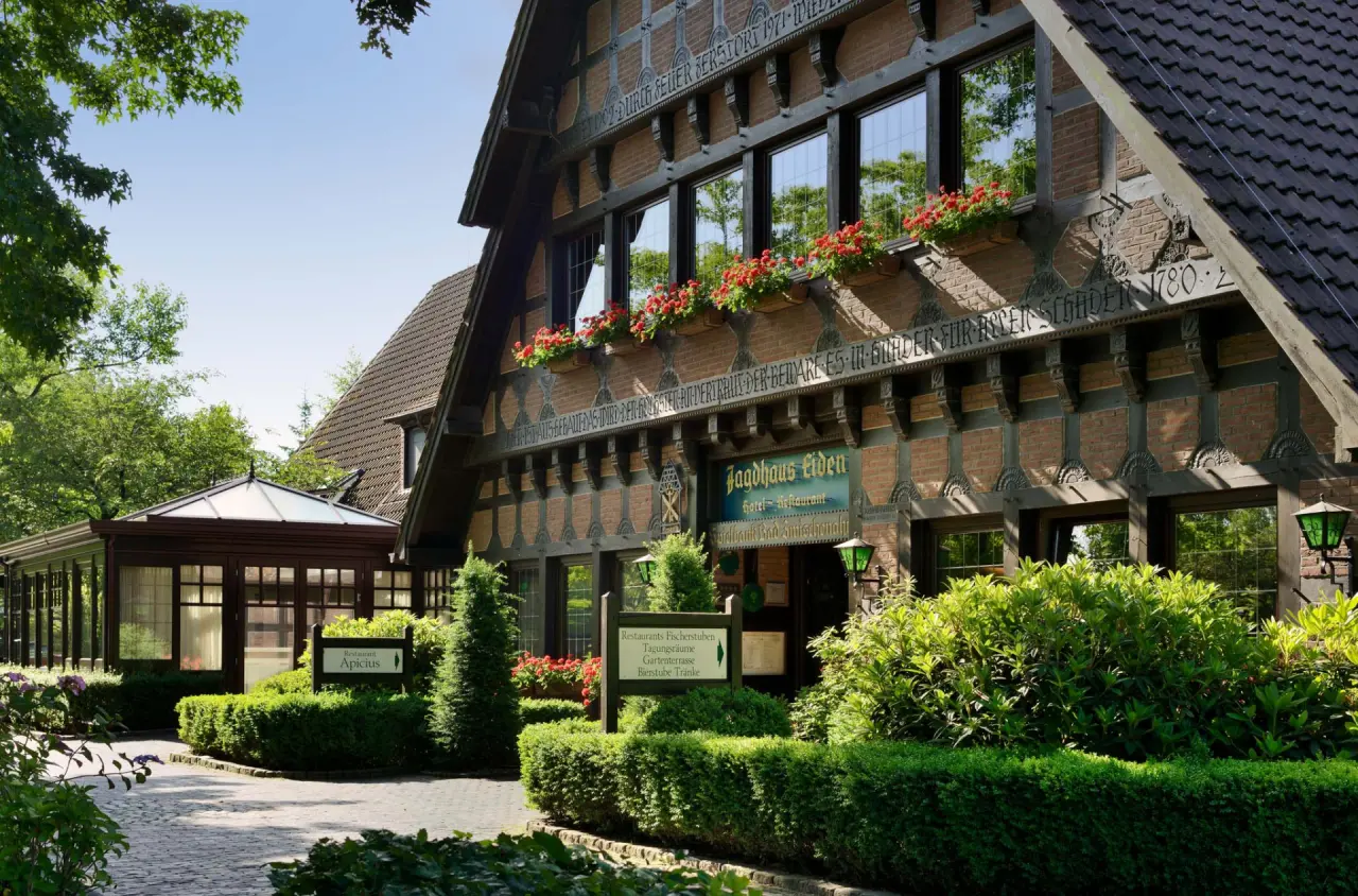 Romantik Hotel Jagdhaus Eiden: Traditionelle Fachwerkfassade mit roten Blumen und gepflegter Gartenanlage bei Sonnenschein.