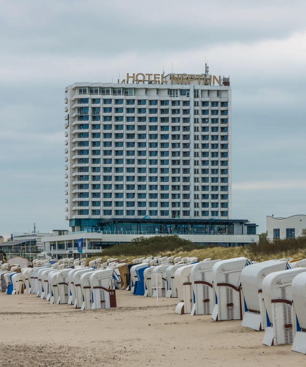 Aussenansicht des Hotel Neptun in Warnemünde mit Strandkörben am sandigen Strand unter bewölktem Himmel.