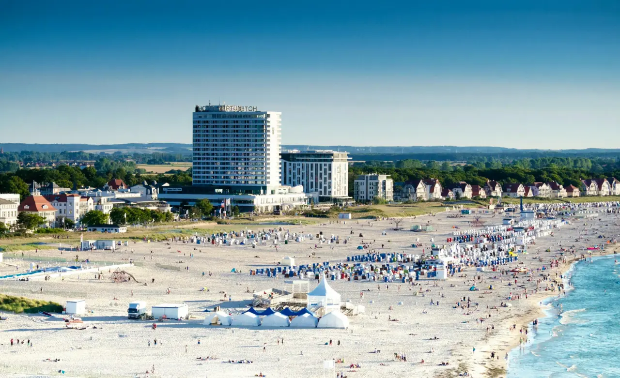 Luftaufnahme des Hotel Neptun an der Ostseeküste mit belebtem Sandstrand und blauem Meer unter strahlendem Himmel.