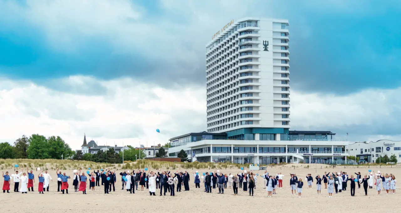 Hotel Neptun an der Ostsee mit Blick auf den Strand und das Meer. Mitarbeiter winken vor dem modernen Hotelgebäude.